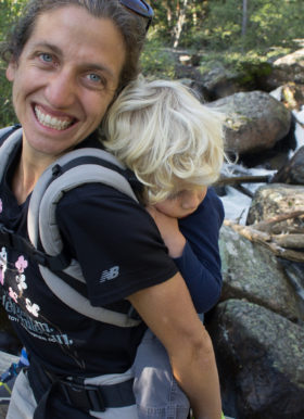 Woman hiking with small blond child on her back and steam behind her